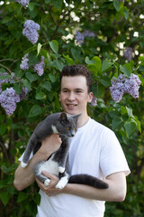 A young man holding a gray cat by a lilac bush.