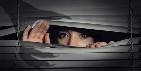 Worried woman looking through window blinds into darkness. Shadow of hand with long claws. Paranoia...