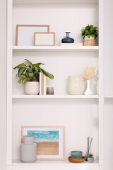 Fototapeta na wymiar Interior design. Shelves with stylish accessories, potted plants and frames near white wall