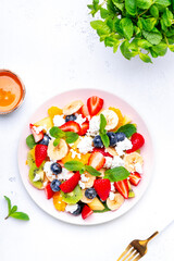 Summer fruit and berry salad with fresh strawberries, blueberries, banana, cottage cheese and mint on white table background, top view