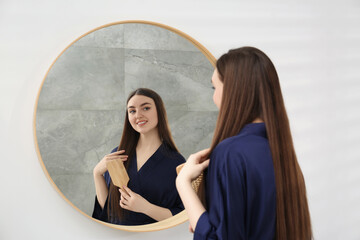 Beautiful woman in blue robe brushing hair near mirror indoors