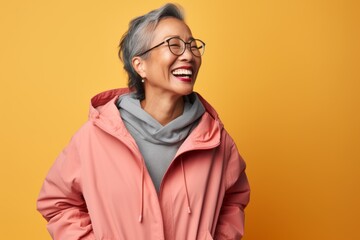 Fototapeta premium Portrait of happy mature Asian woman in pink coat and eyeglasses laughing against yellow background