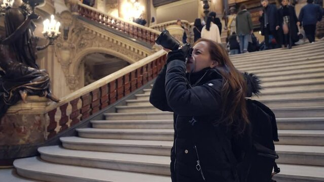 Young Hispanic woman in warm outerwear using photo camera while exploring amazing interior of opera house Palais Garnier with baroque decorated in Paris, France