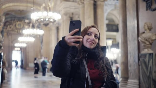 Young Hispanic woman in warm outerwear using photo camera while exploring amazing interior of opera house Palais Garnier with baroque decorated in Paris, France