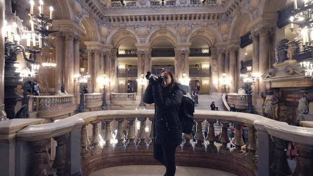 Young Hispanic woman in warm outerwear using photo camera while exploring amazing interior of opera house Palais Garnier with baroque decorated in Paris, France