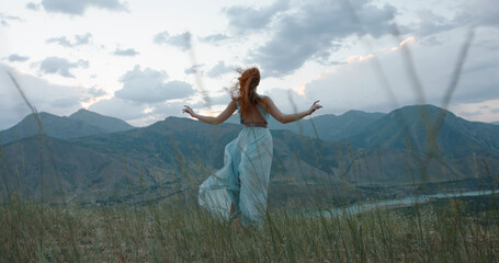 Beautiful carefree girl with red hair wearing white dress standing against the wind, looking at mountains and raising her hands - freedom, adventure, inspiration