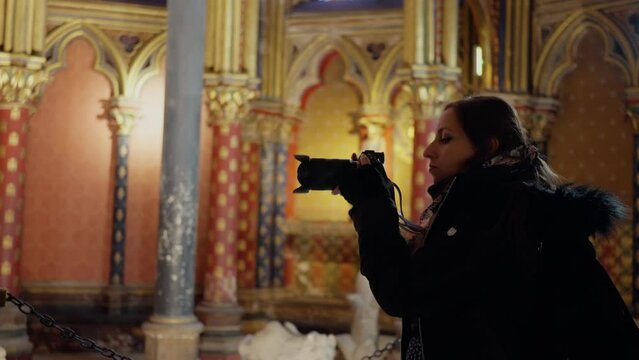 Side view of young Hispanic woman in warm jacket taking photo with professional camera of majestic interior of the Gothic chapel Sainte Chapelle in city of Paris, France