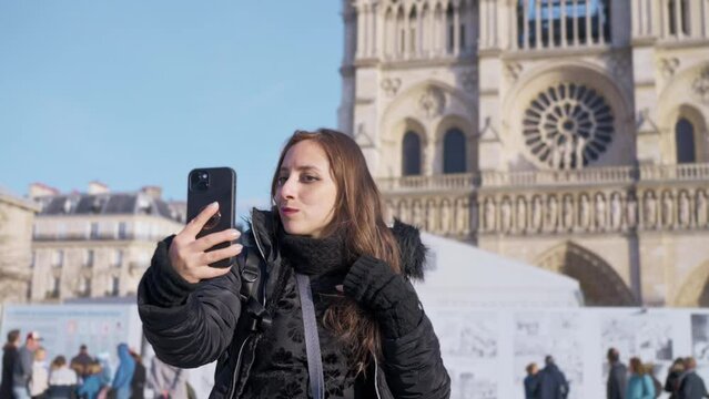 Positive Latina woman tourist smiling and taking selfie on smartphone while standing against landmark of Paris, France in Notre Dame cathedral