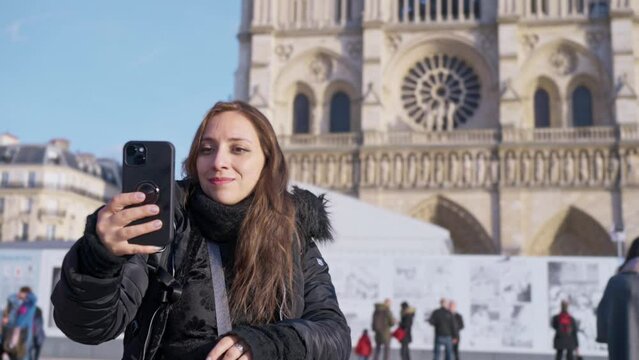 Positive Latina woman tourist smiling and taking selfie on smartphone while standing against landmark of Paris, France in Notre Dame cathedral