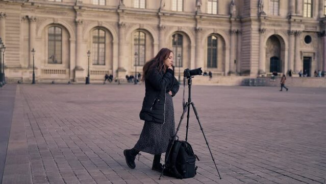 Positive Latina woman photographer in warm clothes taking photos on professional photo camera on tripod or stationary holder while standing near Louvre museum in Paris, France