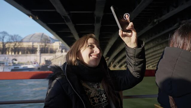 Smiling young Latina woman in warm clothes taking photos while having boat trip on river Seine with aged bridge in Paris, France