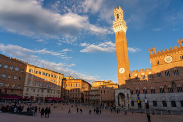 Fototapeta na wymiar Sunset in the Piazza del Campo square of Siena, Italy. The Torre del Mangia is bathed in golden light, as well as the tops of some buildings. People visible on the town square.