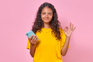 Young optimistic brunette Indian woman with phone in hand shows OK gesture as sign of agreement with your opinion or endorsing proposed plan for weekend stands on isolated pink background.
