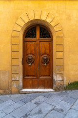 Medieval brown wooden door with two lion head doorknobs set inside a buttercream yellow colored arched doorway and wall in the city of Siena, Italy. 