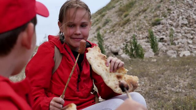 Two children brother and sister eat fried sausages with bread on a fire at a picnic in the mountains. Teenagers love barbecue while hiking in the mountains. Hot sausages fried on the fire by children
