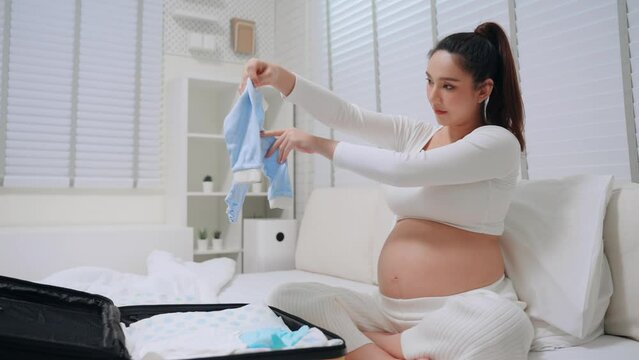 Asian pregnant woman arranging pack necessities and baby items in luggage at home preparing prenatal. Maternity prenatal care and pregnancy concept.