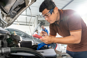 An Asian male car mechanic using his phone while working on repairing a car's engine at car repair...