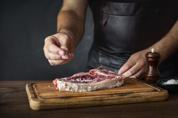 Man cook in apron sprinkles salt on a fresh raw beef steak on the bone