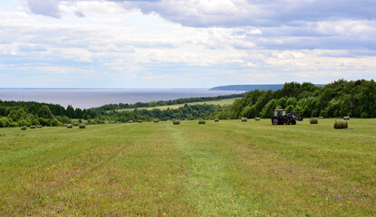 agricultural field with bales of hay and working tractor with forest line and river  