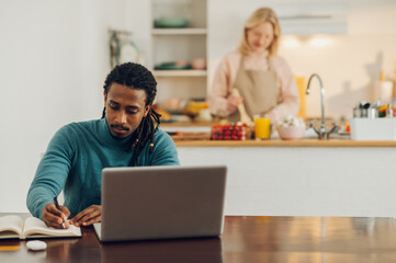 A multicultural casual entrepreneur is sitting at a table with a laptop and working while his wife is cooking in the kitchen.