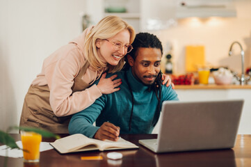 A cheerful housewife is hugging her busy interracial man while standing at home.