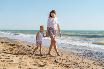 young woman walks with her son on seashore in t-shirt and shorts, walk along shore at sunset, vacation concept