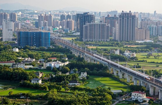 A train of High Speed Rail traveling on a viaduct thru green rice fields with houses scattered in the farmlands and high rise buildings booming in background, in Zhubei City, Hsinchu County, Taiwan