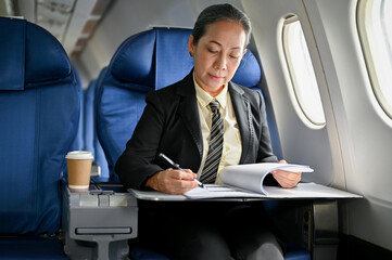 A successful mature Asian businesswoman focuses on reviewing business reports during the flight