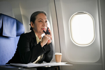 A thoughtful senior Asian businesswoman is on the phone with her client during the flight.