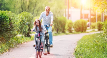 Schilderijen op glas Portraits Smiling father with daughter during summer outdoor bicycle riding. They enjoy togetherness in the summer city park. Happy parenthood and childhood or active sport life concept image. © Soloviova Liudmyla
