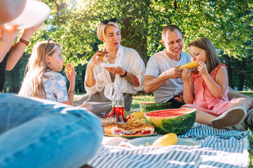 Big family sitting on the picnic blanket in city park during weekend Sunday sunny day. They are smiling, laughing and eating boiled corn and watermelon. Family values and outdoors activities concept.