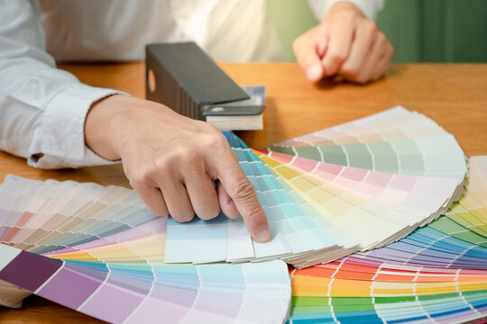 Designer hand choosing and pointing at color swatch samples catalog or rainbow colour palette guide for selection coloring on architecture and interior design work.