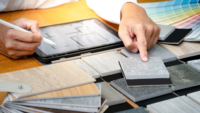 Architect hand choosing and pointing stone and wood material samples while working with digital tablet on the table in studio. Designer working for interior architecture and furniture design project.