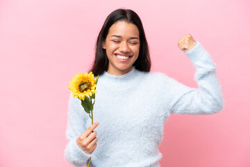 Young Colombian woman holding sunflower isolated on pink background doing strong gesture