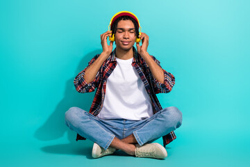 Full length photo of peaceful satisfied young man sitting floor closed eyes arms touch headphones isolated on teal color background