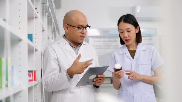 Handsome asian male pharmacist and coworker wearing lab coat and checking medicine product stock with database tablet in the pharmacy drugstore. 4k resolution.