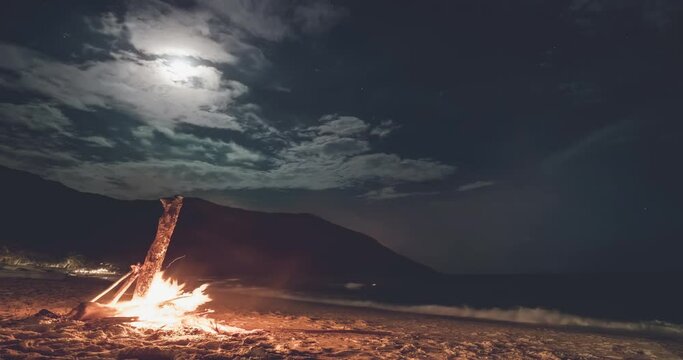 Beach bonfire at night under moonlight. Cloud timelapse above mountain island. Burning wood on sand shore. Dark sky with stars. Tropical romantic landscape near ocean with waves. Camp atmosphere.