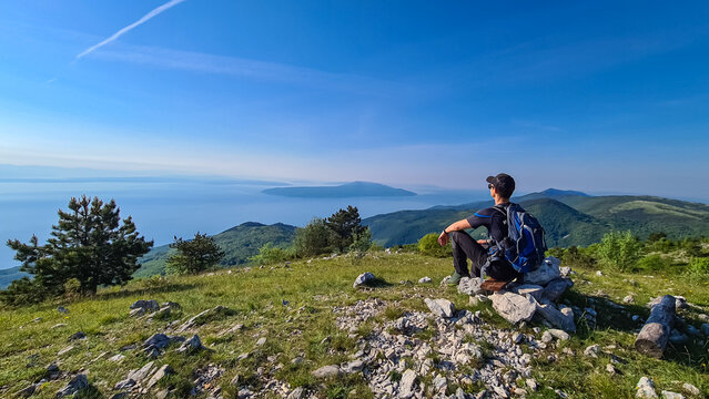 A man sitting on top of Vojak and enjoying the panoramic view on Mediterranean Sea. The mountain is overgrown with lush green plants. Few islands in the back. Early morning hiking by the sea. Remedy