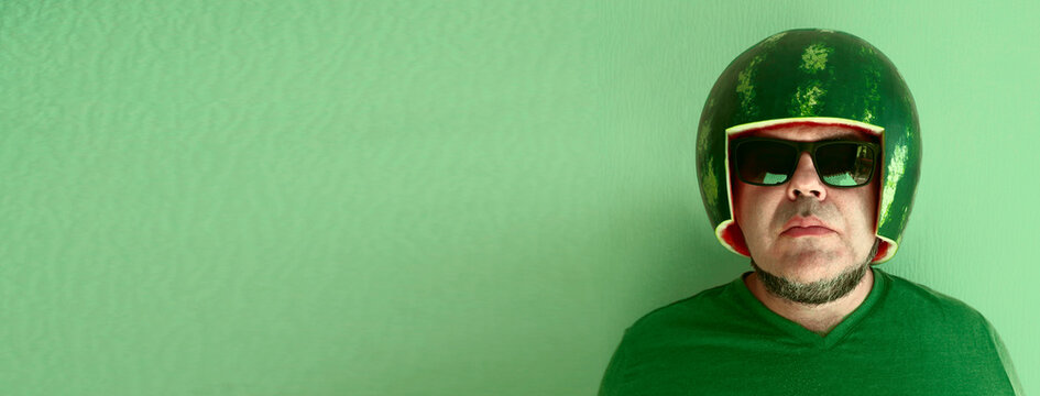 Crazy man wearing watermelon as helmet, hard hat on head and stylish sunglasses. male isolated on green wallpaper background. idea of protection and insurance