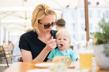 Young caucasian blonde mother spoon feeding her little infant baby boy child outdoors on restaurant...