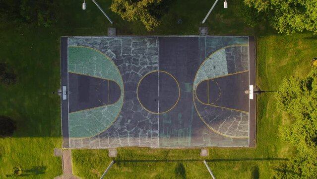 Aerial Drone View Of Cracked Concrete Empty Basketball Court In Outdoor Park Surrounded By Lush Green Grass And Trees During Sunny Sunrise Sunset.