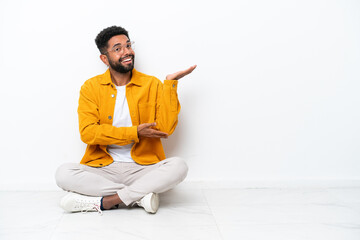 Young Brazilian man sitting on the floor isolated on white background extending hands to the side for inviting to come - obrazy, fototapety, plakaty