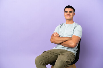 Young man sitting on a chair over isolated background keeping the arms crossed in frontal position