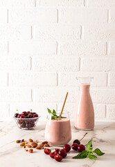 A freshly made homemade smoothie made with ripe cherries in a large glass glass with almonds and mint and a full bottle of smoothie. White brick wall.