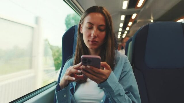 Cute smiling girl with long brown hair, dressed in blue shirt, while traveling by suburban train, scrolls on smartphone screen