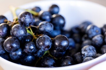 A portrait full of fresh multiple black or blue grapes in a white bowl. The fruit bunch is healthy and a great small snack to eat during a break or during a party with friends or family.