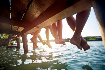 Close-up shot of a barefoot group of friends sitting on the dock on the river. Summer, river, vacation
