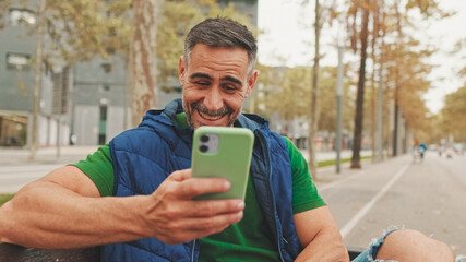 Mature man wearing casual clothes, sitting on bench in the park, talking on video call from smartphone