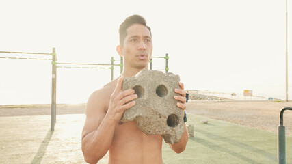 Guy, the athlete focused on lifting a stone during a workout, does an exercise of the muscles on an outdoor sports field on a summer day