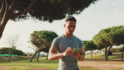 Guy in sportswear, an athlete walks through a city park, scrolls on the screen of a mobile phone, types a message on a smartphone